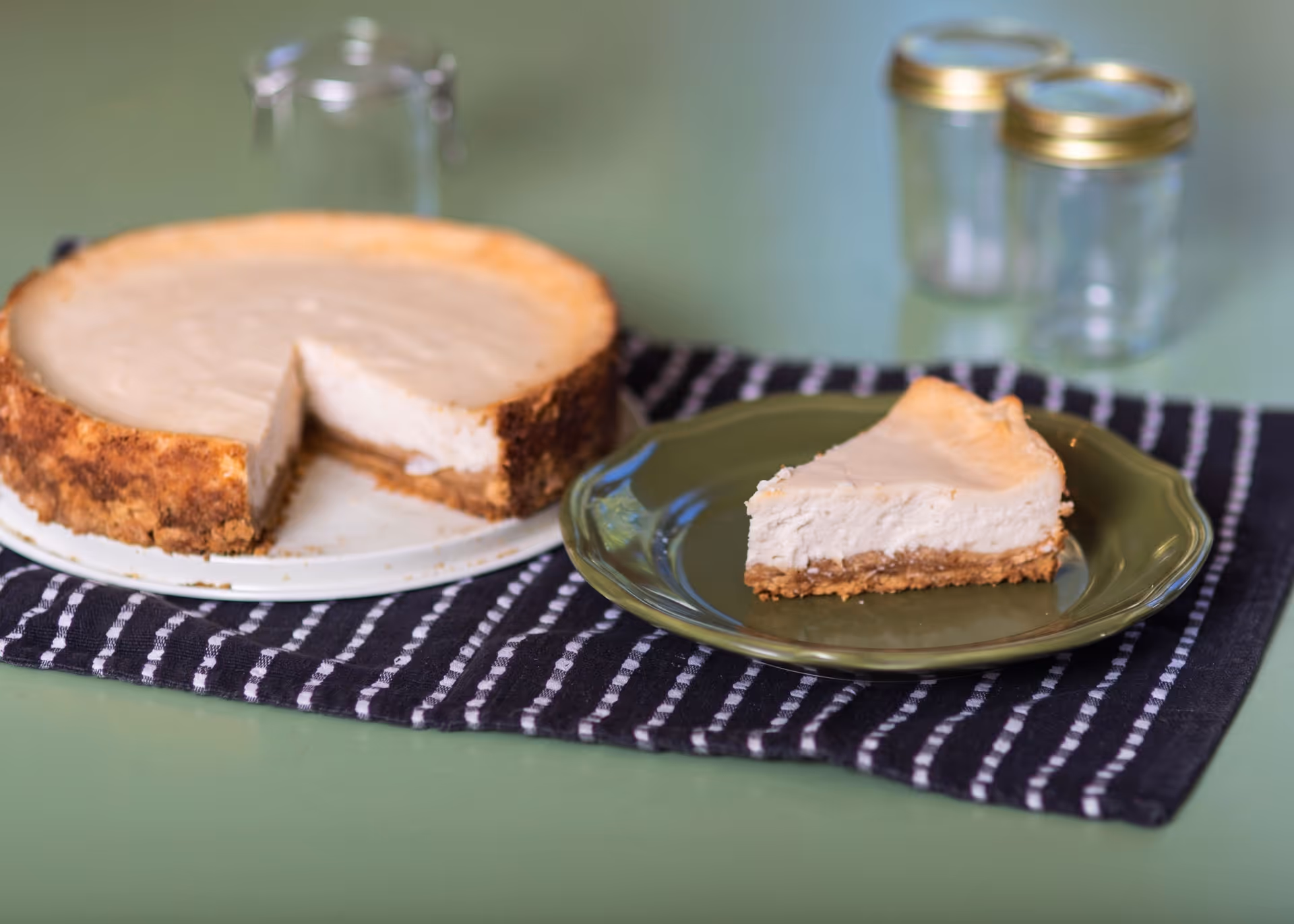 A slice of cheesecake with a graham cracker crust on a green plate beside the remaining cheesecake on a white tray.