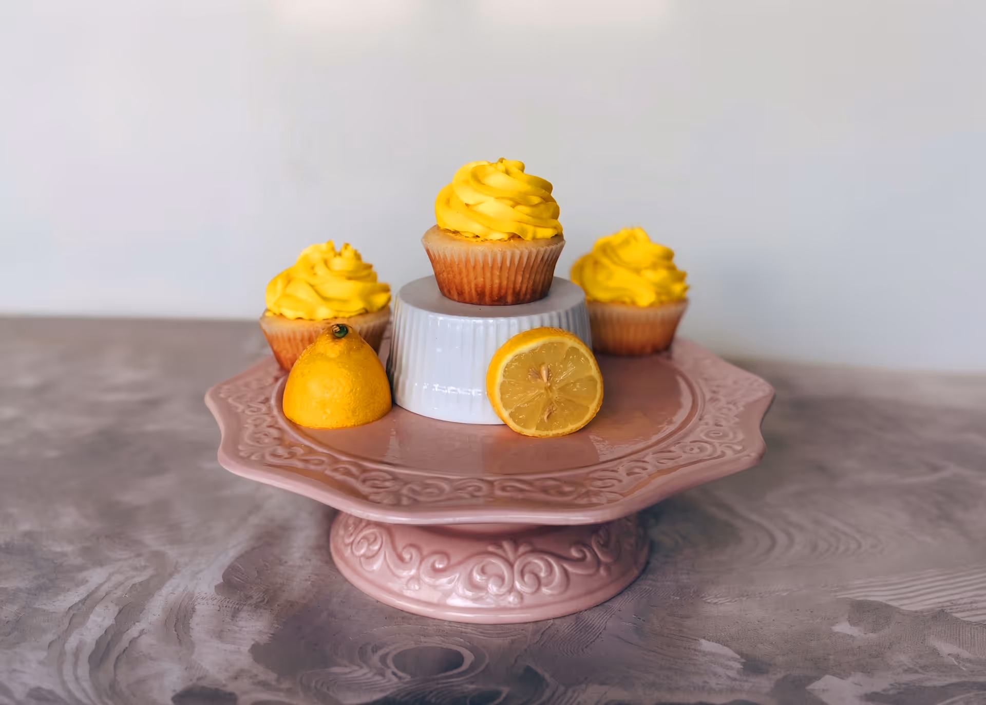 Three lemon cupcakes with yellow frosting arranged on a pink decorative cake stand with a white small stand in the center and two lemon halves.
