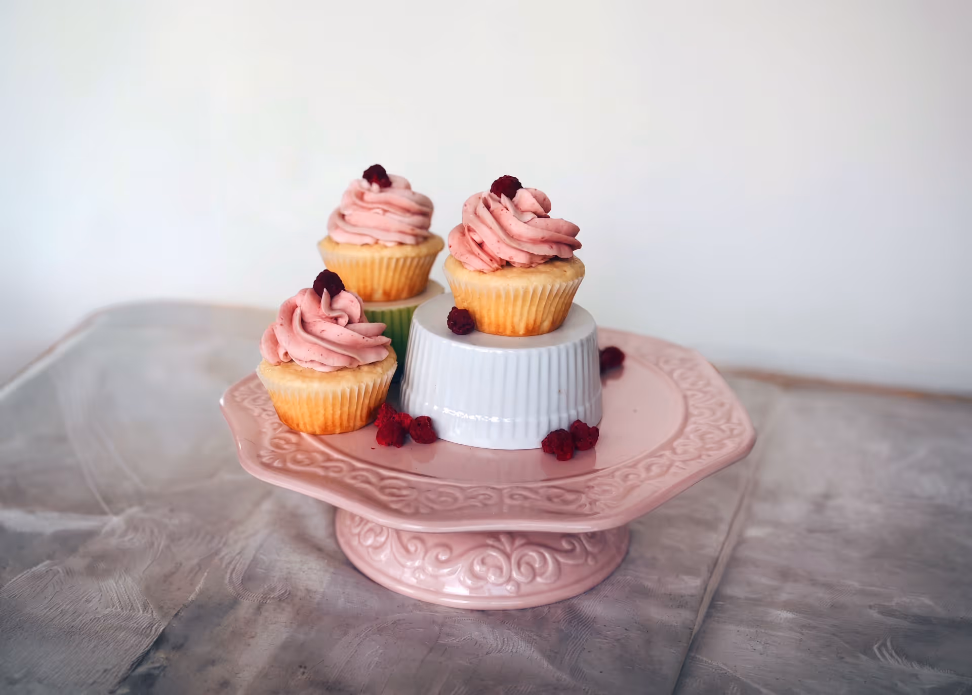 Three vanilla cupcakes with pink frosting, topped with a berry on a pink decorative cake stand.