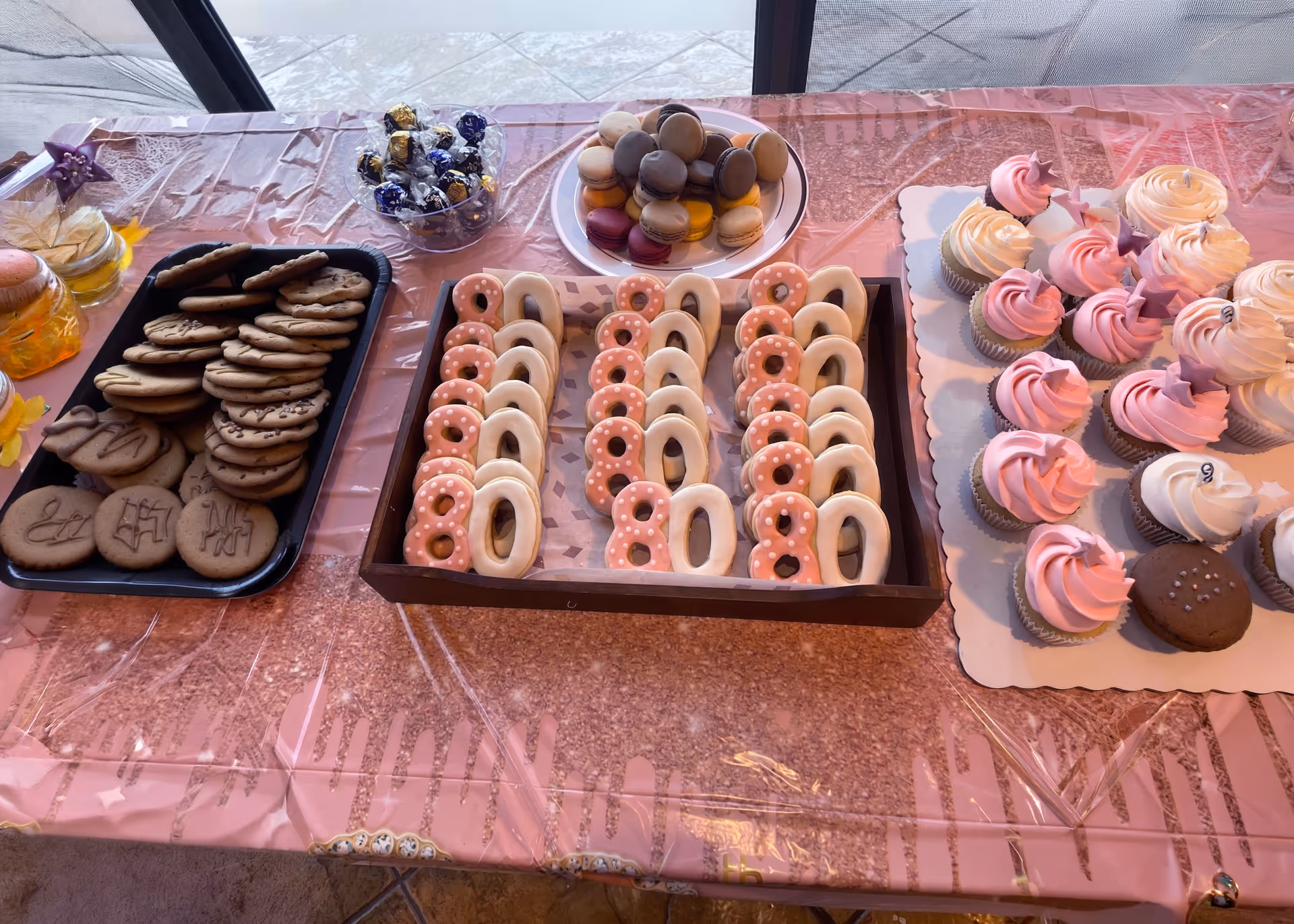 Table with trays of assorted cookies, decorated pink and white number 80 cookies, colorful macarons, and pink and white frosted cupcakes with star toppers.