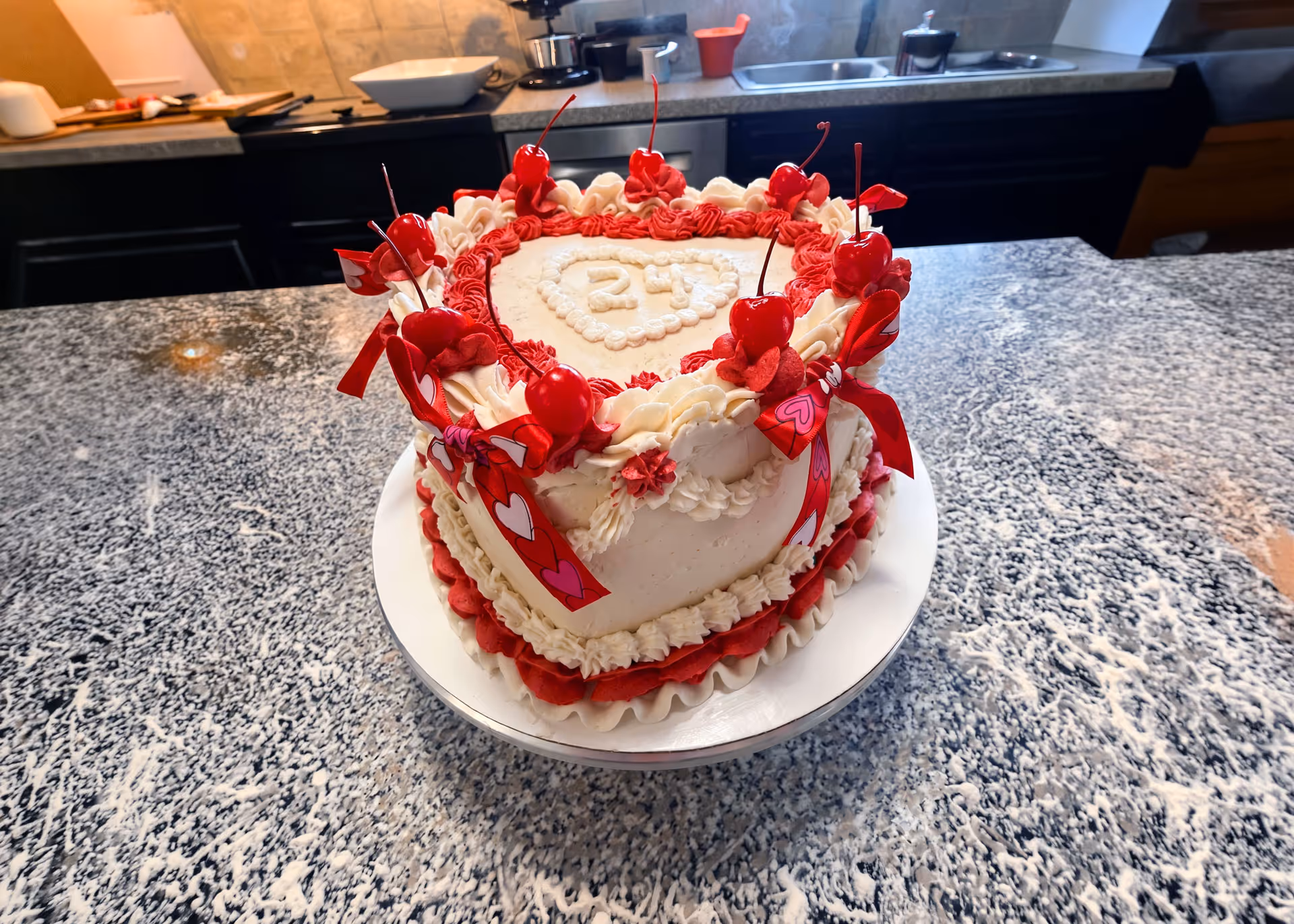 Heart-shaped cake with red and white frosting, decorated with cherries and red bows on a kitchen countertop.