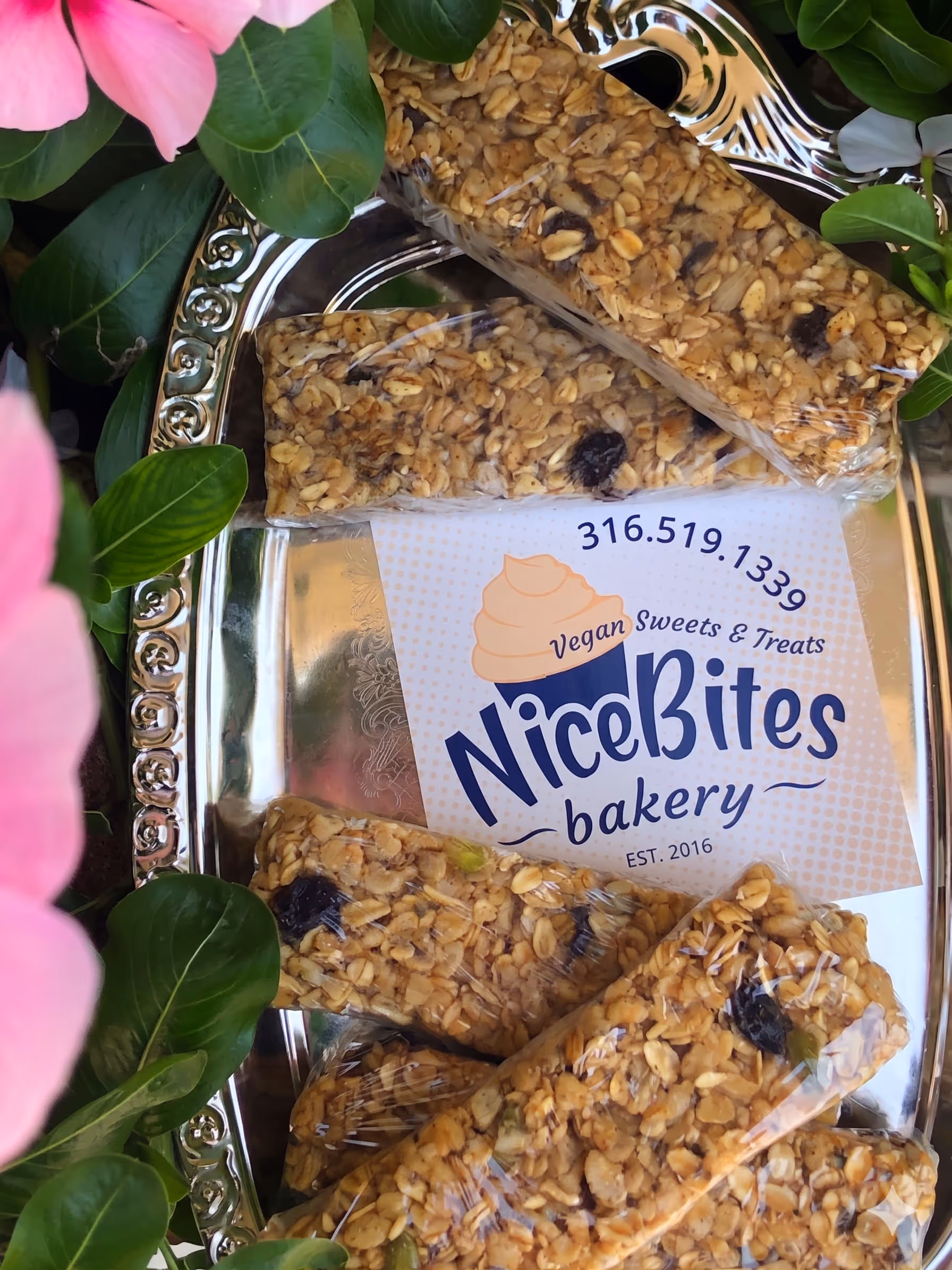 Granola bars wrapped in plastic on a silver tray with NiceBites Vegan Sweets & Treats bakery card surrounded by green leaves and pink flowers.