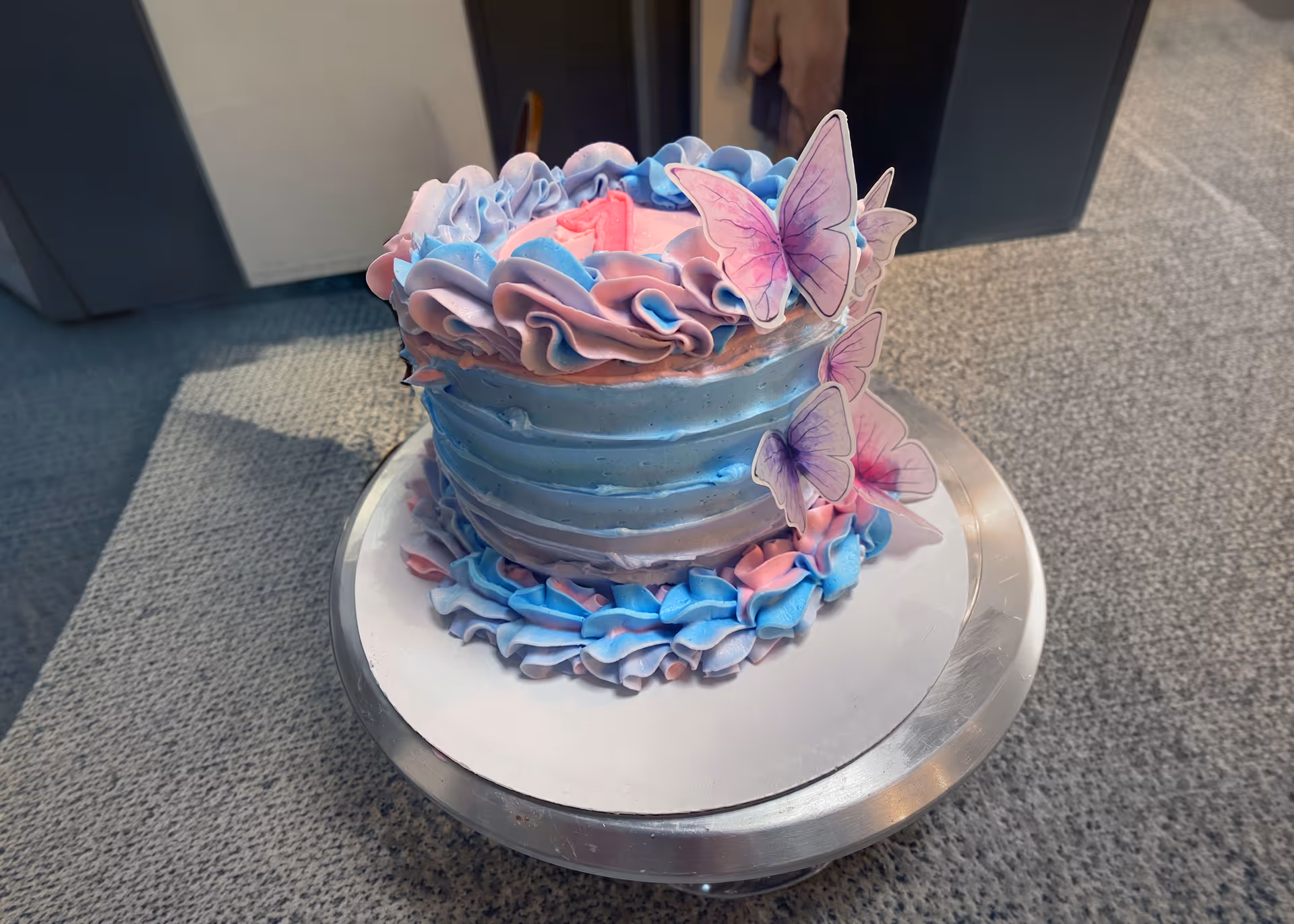 Blue and pink birthday cake decorated with swirled frosting and paper butterflies on a silver cake stand.