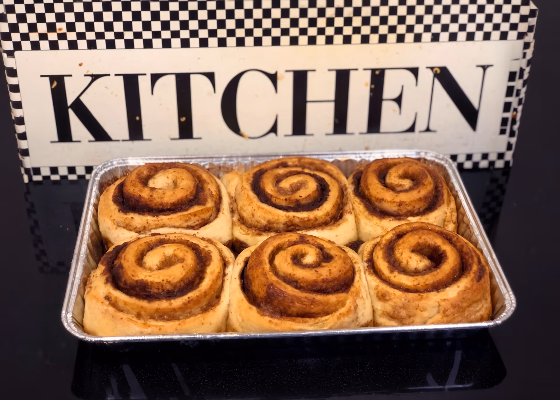 A tray with six cinnamon rolls placed in front of a checkered kitchen sign.