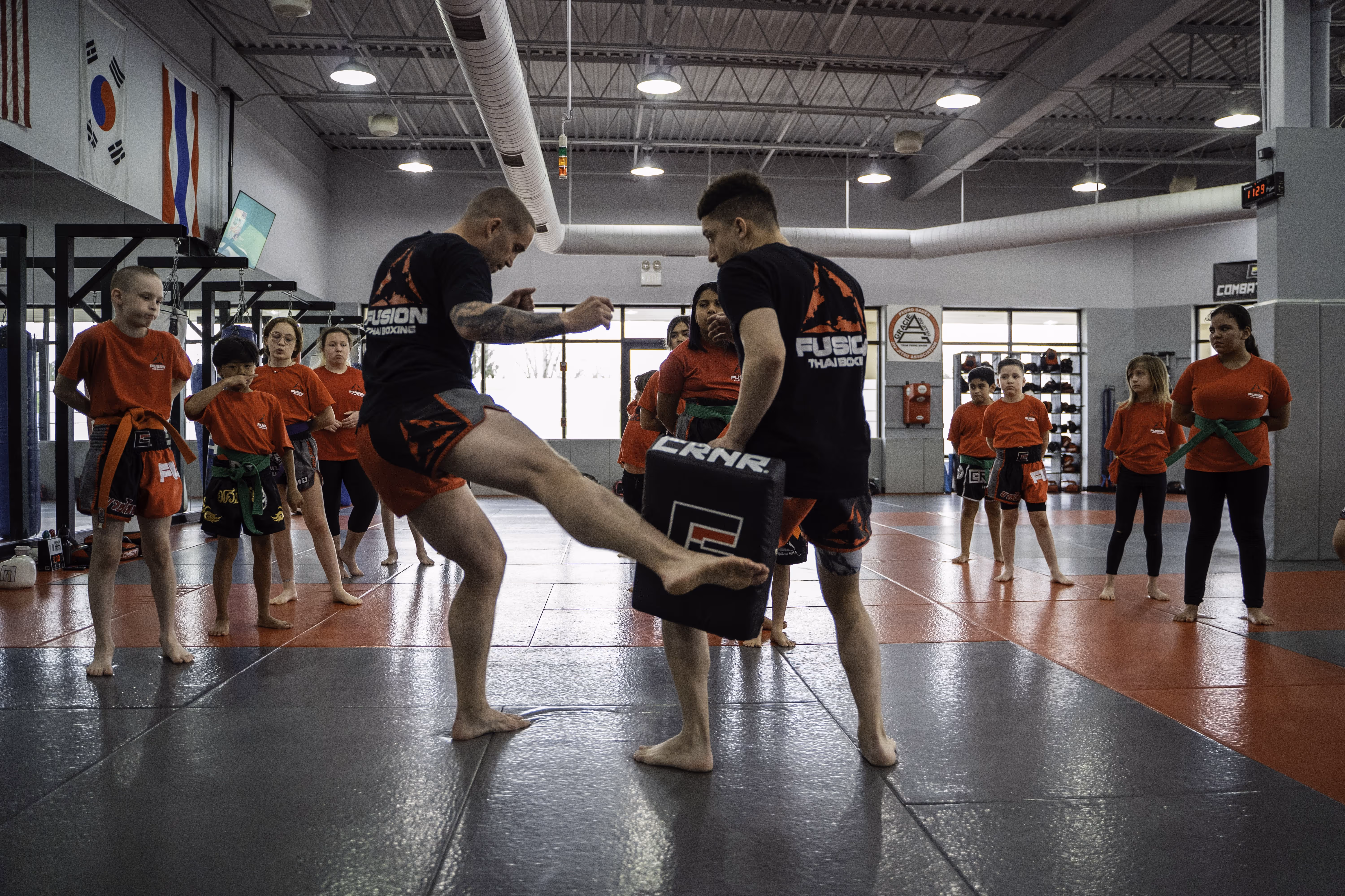 Martial arts class with kids in red shirts watching two instructors practice kicking and holding pads.