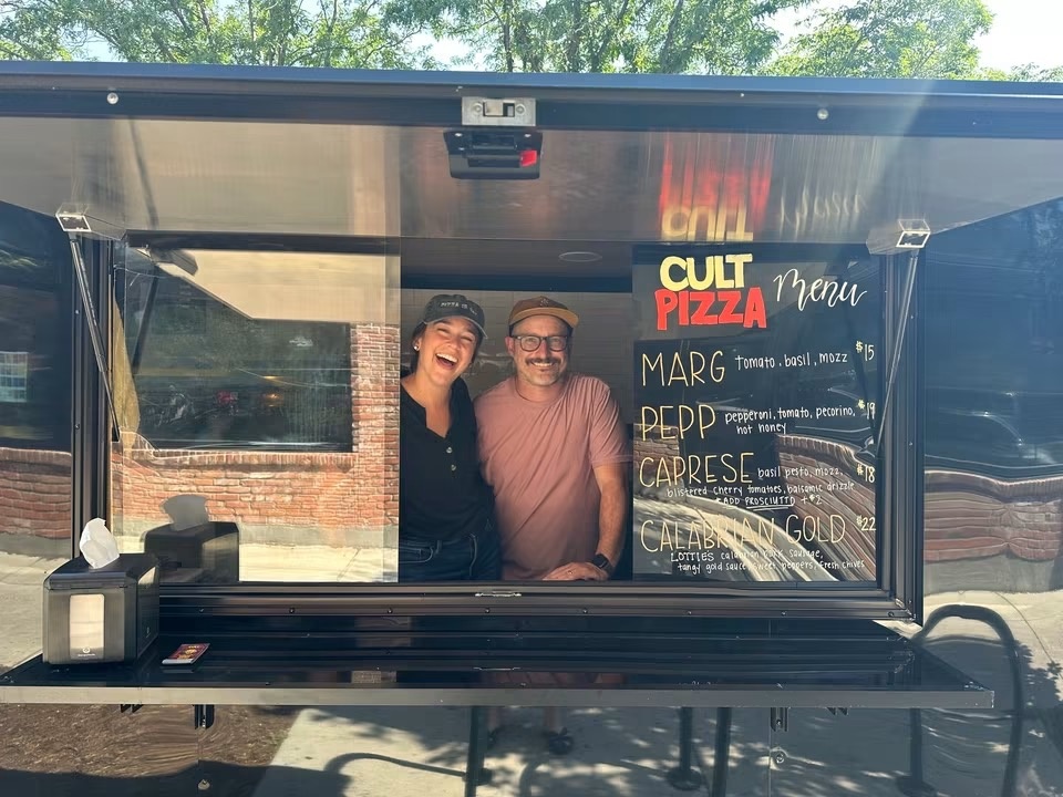 Tim and Paige smiling in the serving window of their Cult Pizza trailer