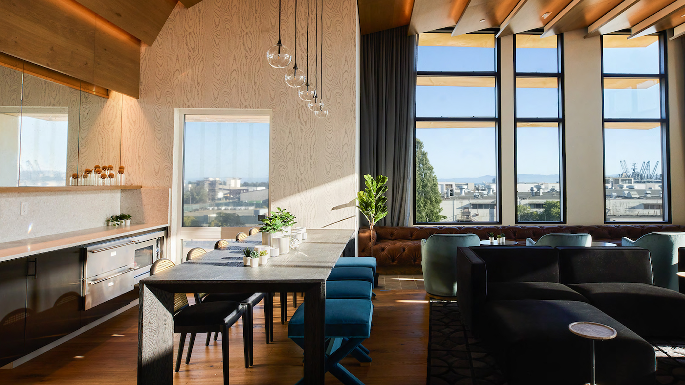 Modern lounge with wooden table, blue stools, black chairs, large windows, and city view.