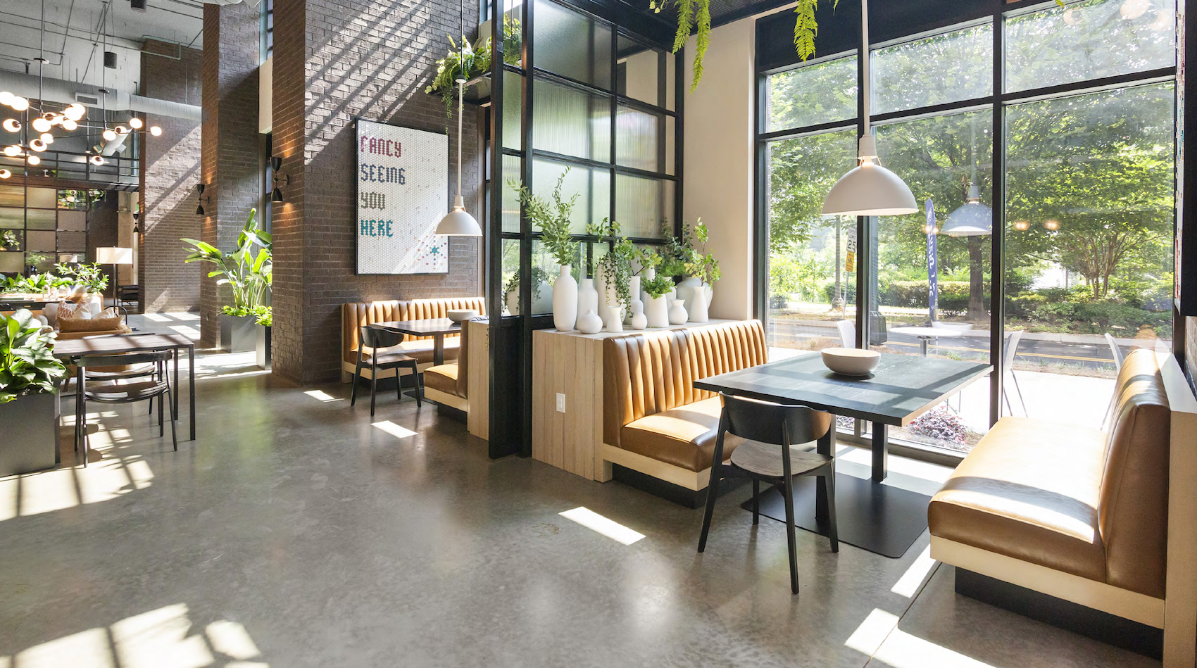 Bright modern cafe interior with tan leather booths, black chairs, white vases with greenery, and large windows.