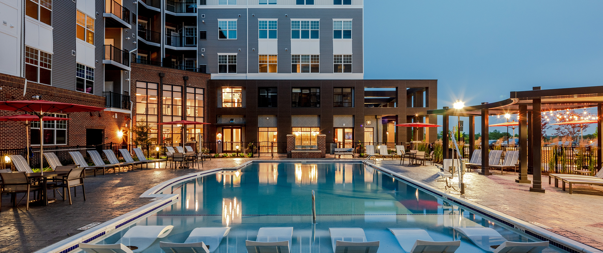 Evening view of a modern outdoor apartment pool area with lounge chairs and umbrellas.