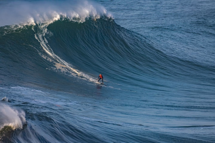 Surfer riding a large blue ocean wave with white spray at the crest.