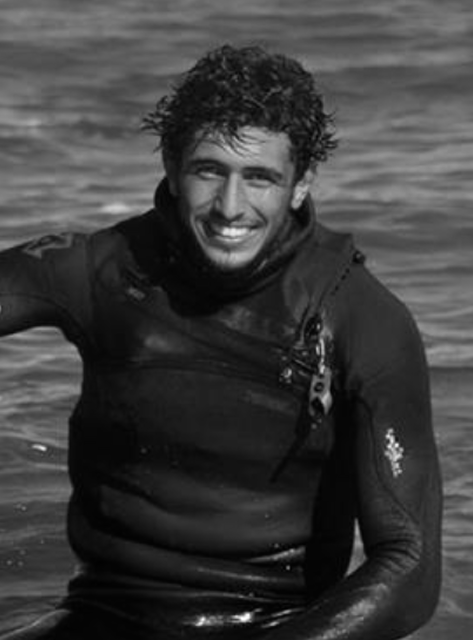 Smiling man with wet curly hair wearing a wetsuit in the water.