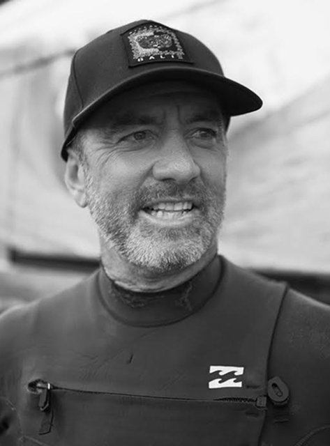 Smiling man with grey beard wearing a cap and wetsuit outdoors.