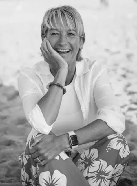 Woman with short hair sitting on sand, smiling, wearing floral pants, white shirt, and smartwatch.