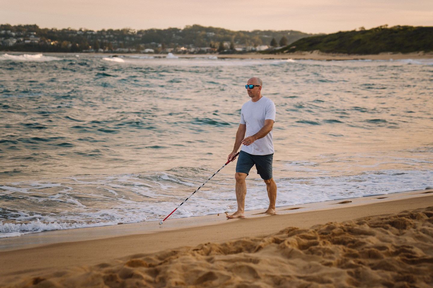 Man with sunglasses walks barefoot on sandy beach near ocean waves using a white cane.