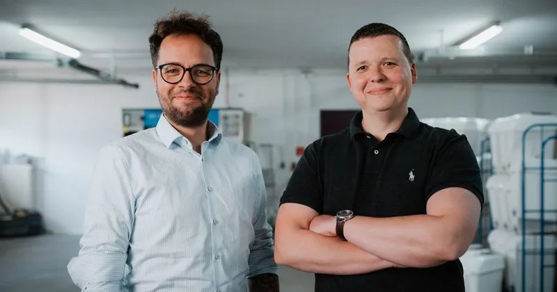 Zwei Männer lächeln in einer hellen Lager- oder Werkstatthalle, einer trägt ein weißes Hemd und Brille, der andere ein schwarzes Polohemd mit verschränkten Armen.
