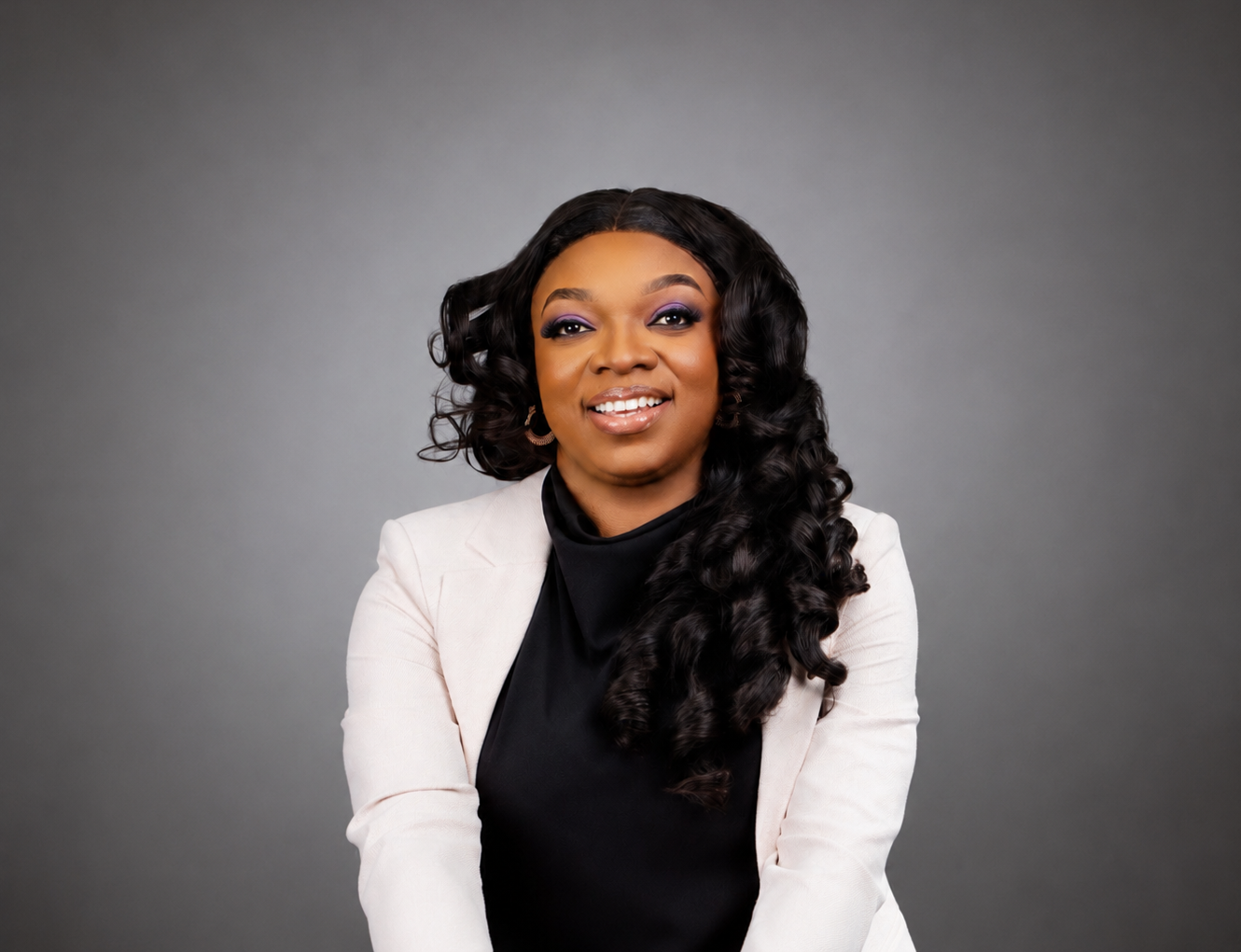 Smiling woman with long curly hair wearing a white blazer and black top against gray background.