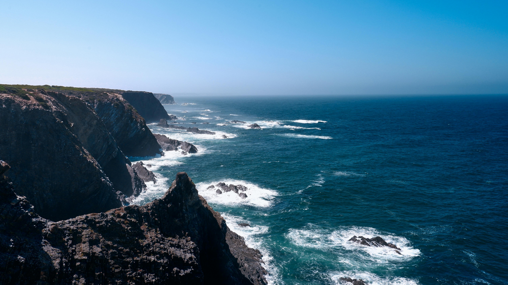 Rocky cliffs along a blue ocean coastline under a clear blue sky.