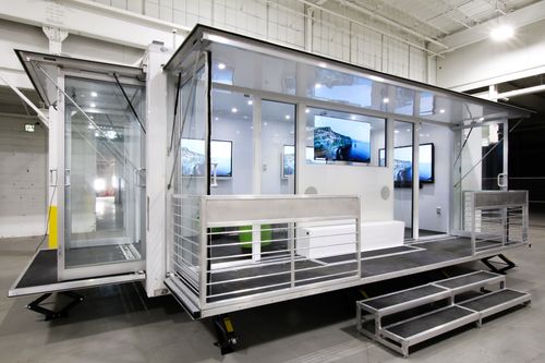 Portable trailer with glass walls, metal railing, steps, and TV screens inside showing coastal images.