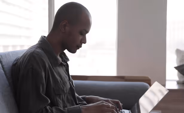 Man focused typing on a laptop while sitting on a couch by a bright window.