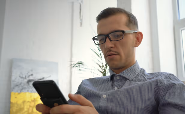 Man with glasses looking intently at a black smartphone while sitting indoors by a window.