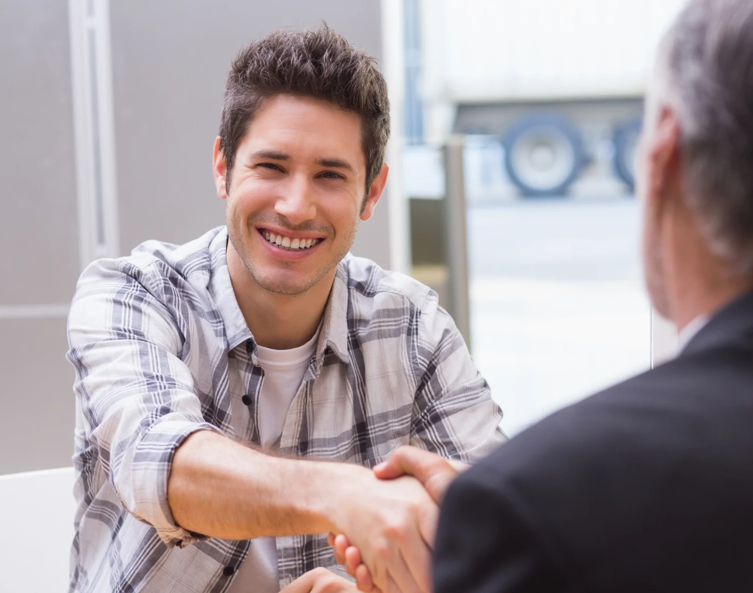 Smiling young man in plaid shirt shaking hands with another person in a business setting.