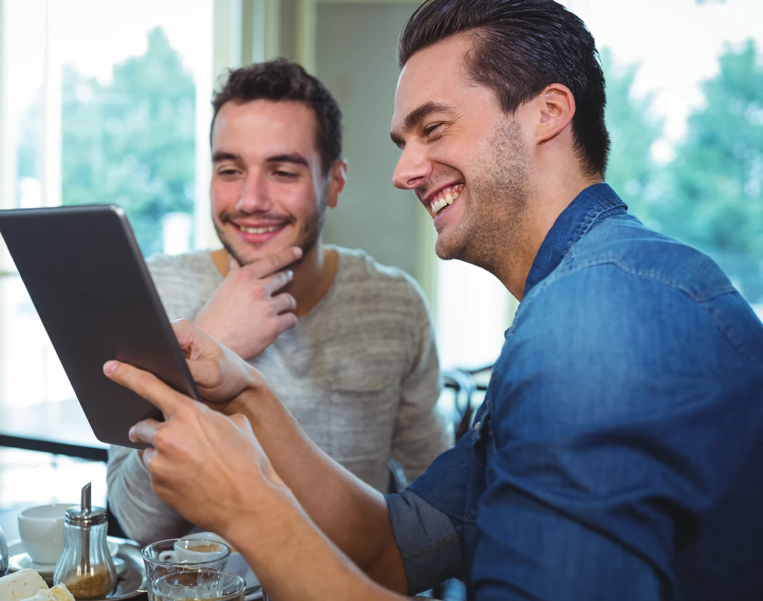 Two men smiling and looking at a tablet together in a bright cafe setting.
