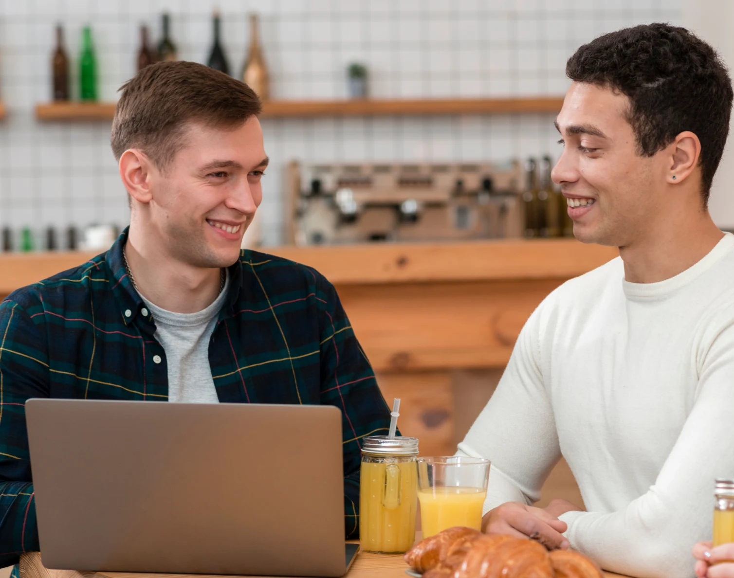 Two men smiling and sitting at a table with a laptop, juice, and croissants in a cafe.