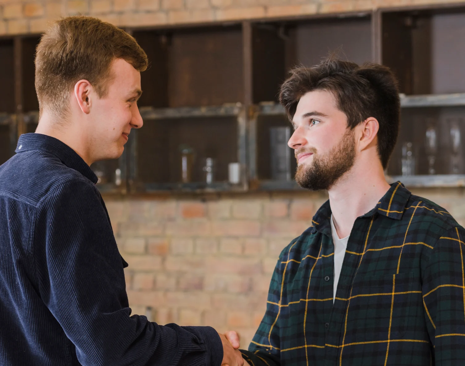 Two young men shaking hands and smiling, standing indoors with a brick wall behind them.