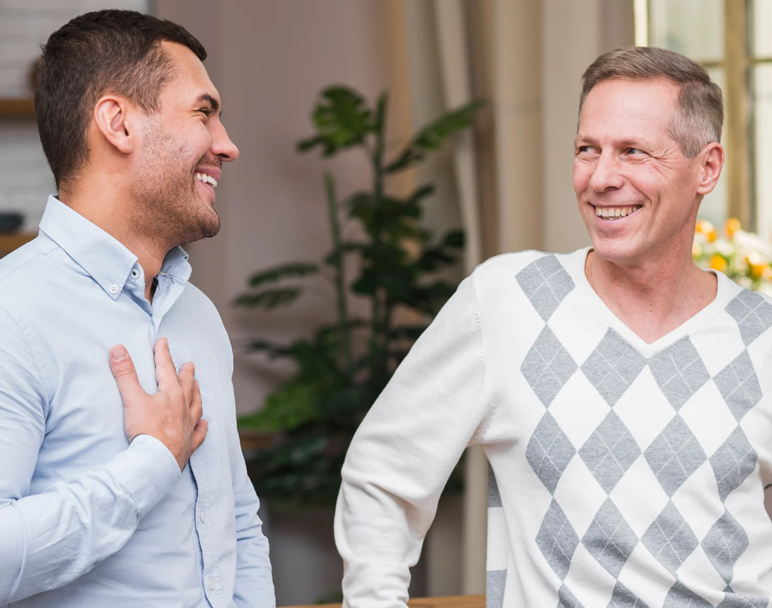 Two men smiling and talking as they stand indoors with plants and curtains in the background.
