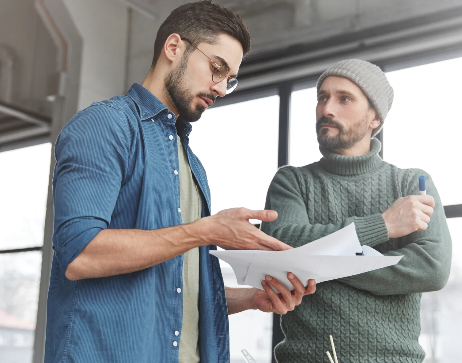 Two men discussing plans indoors, one holding papers and the other with pen and grey knit cap.