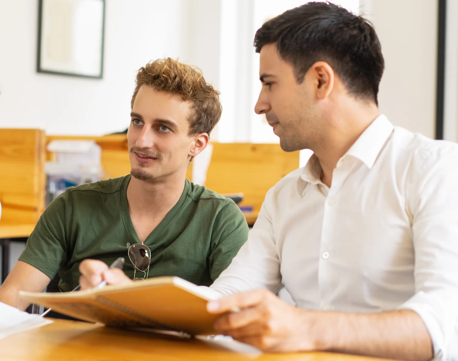 Two men sitting at a table discussing and writing in a notebook indoors.