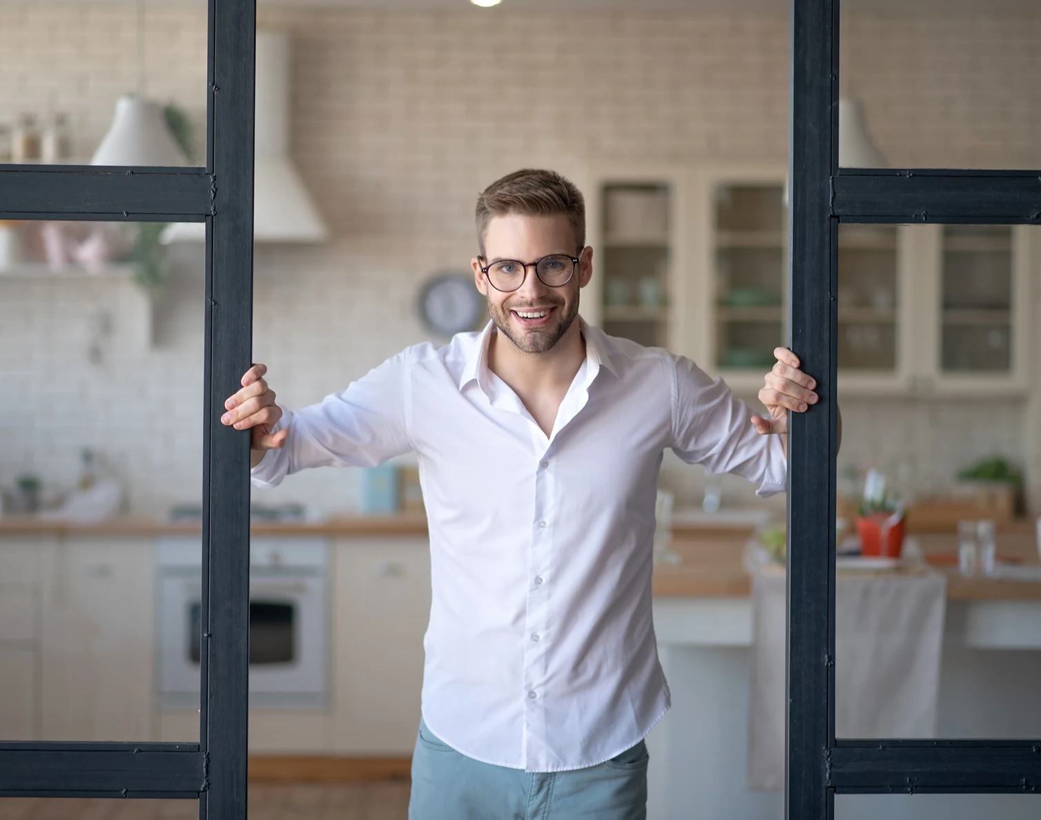 Smiling man with glasses and white shirt standing in a kitchen holding open black-framed doors.