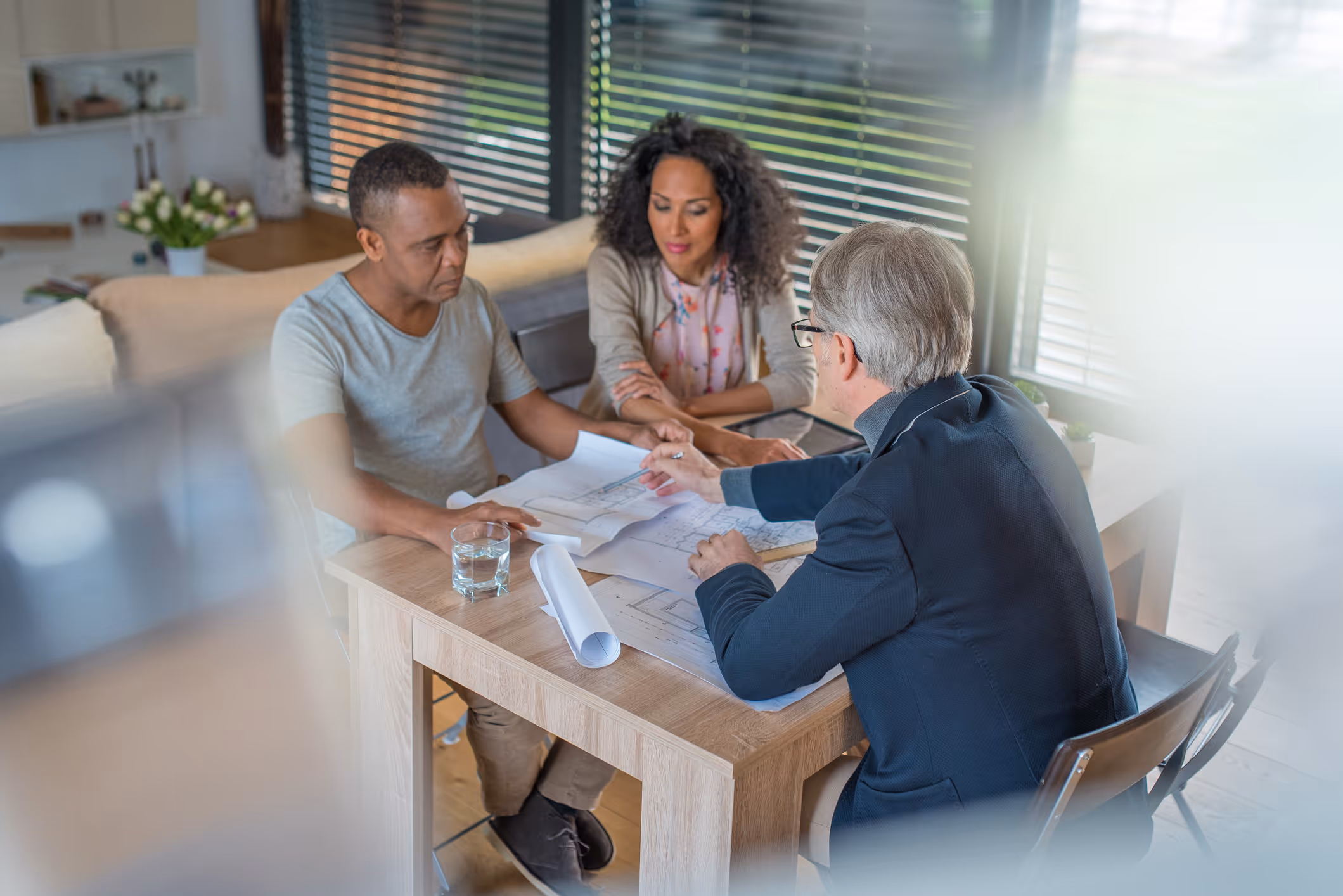 Three people discussing architectural plans at a wooden table by a window with blinds.