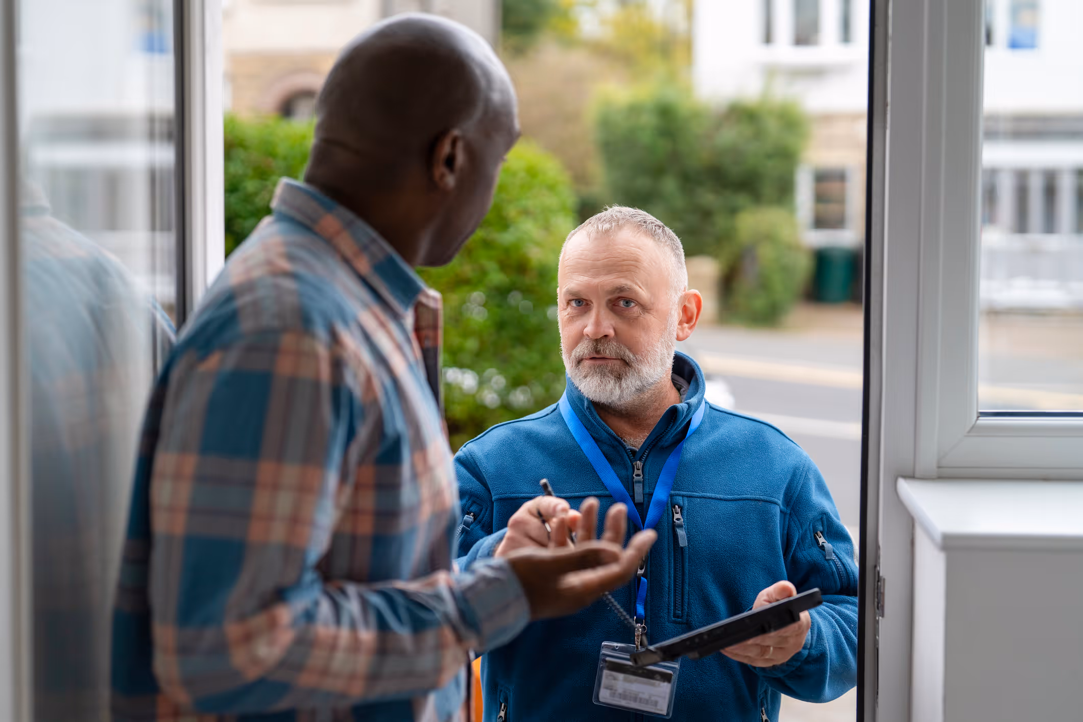 Man in blue jacket with lanyard holding tablet talks to man in plaid shirt at doorway.