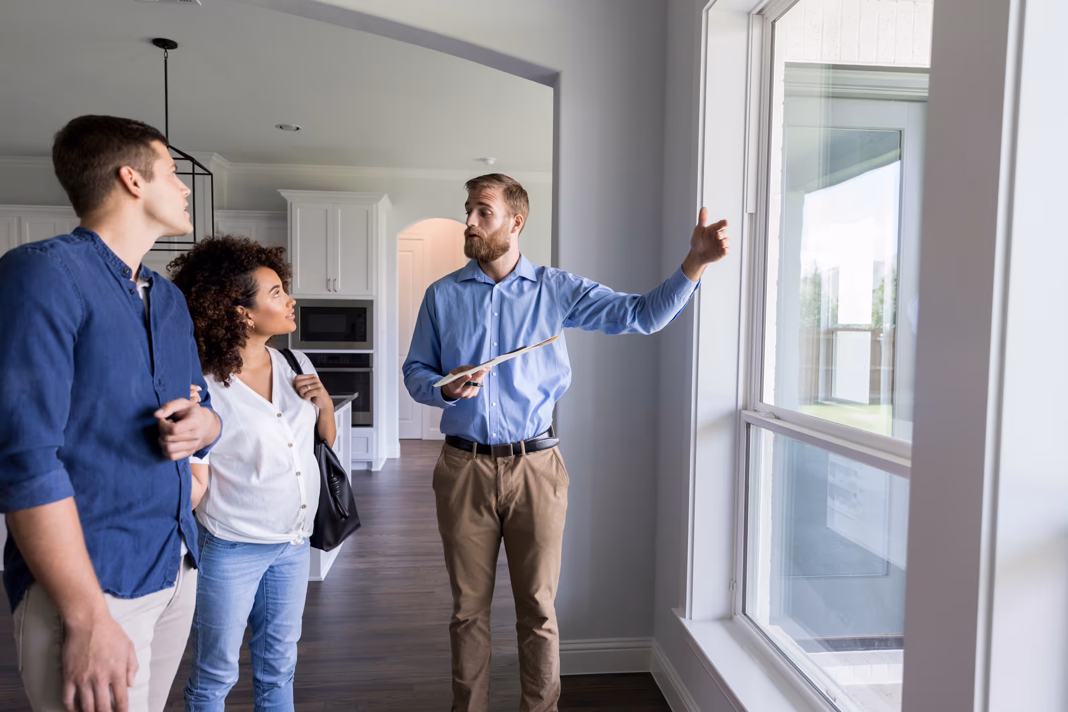 Real estate agent showing a couple a room, pointing toward a large window in a modern home.
