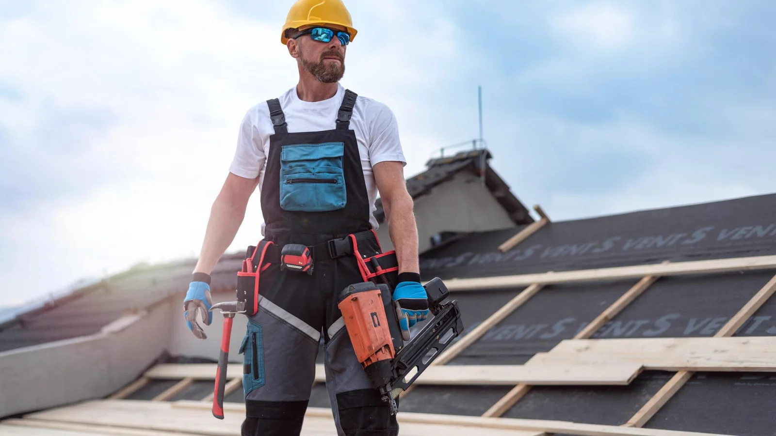 Construction worker wearing helmet and gloves holding a nail gun and hammer on a roof under construction.