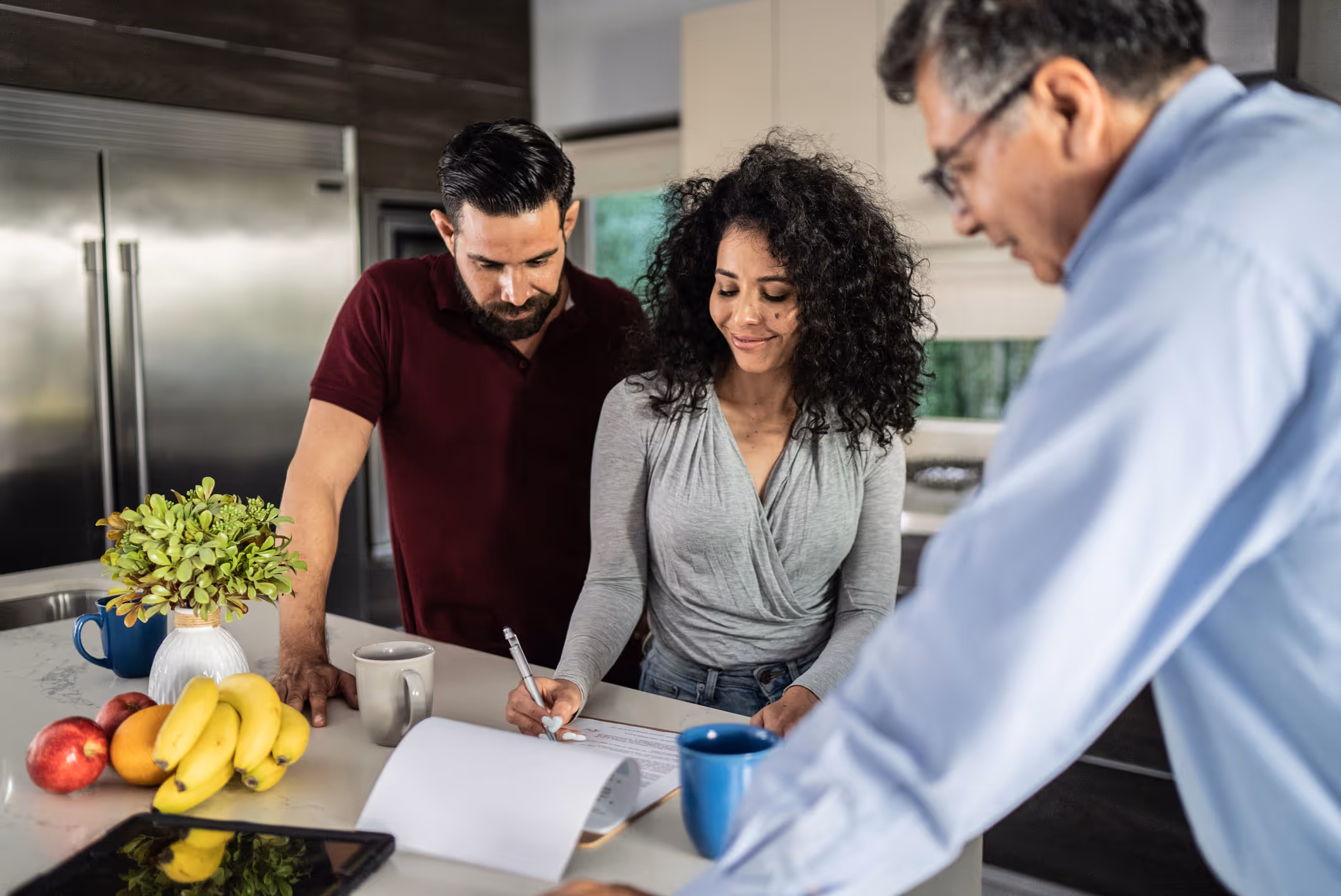 Couple in kitchen reviewing documents with a man helping them, surrounded by fruit and mugs on counter.