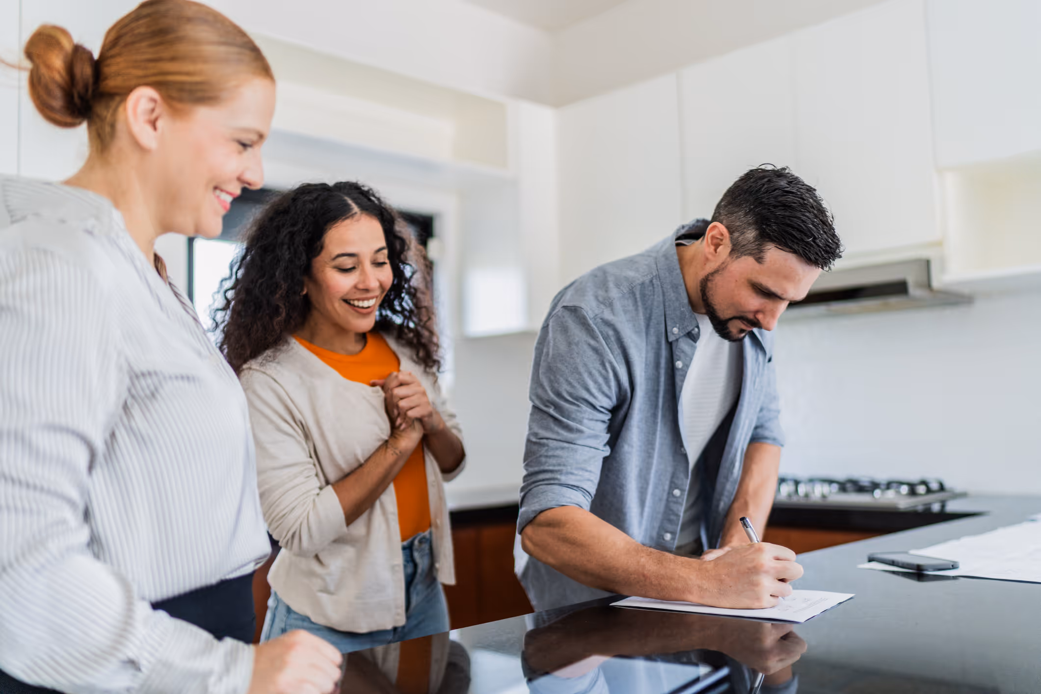 Man signing document on kitchen counter while two women watch and smile nearby.