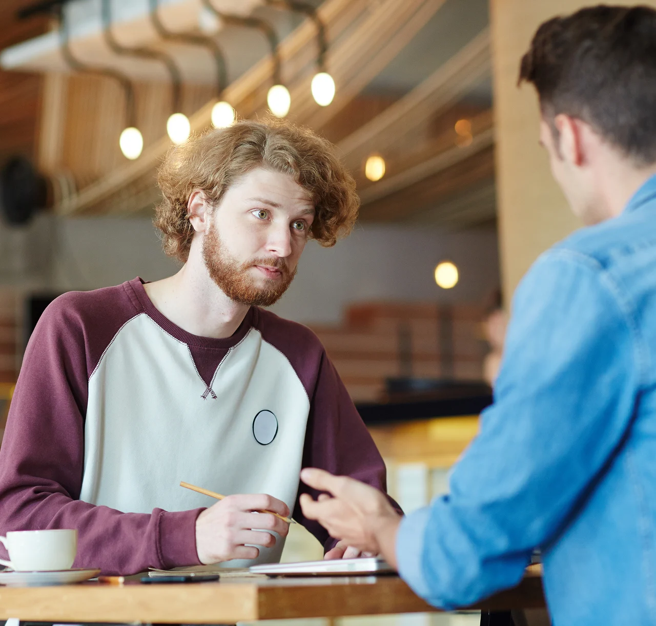 Two men talking at a table in a cafe, one holding a pencil and listening attentively.