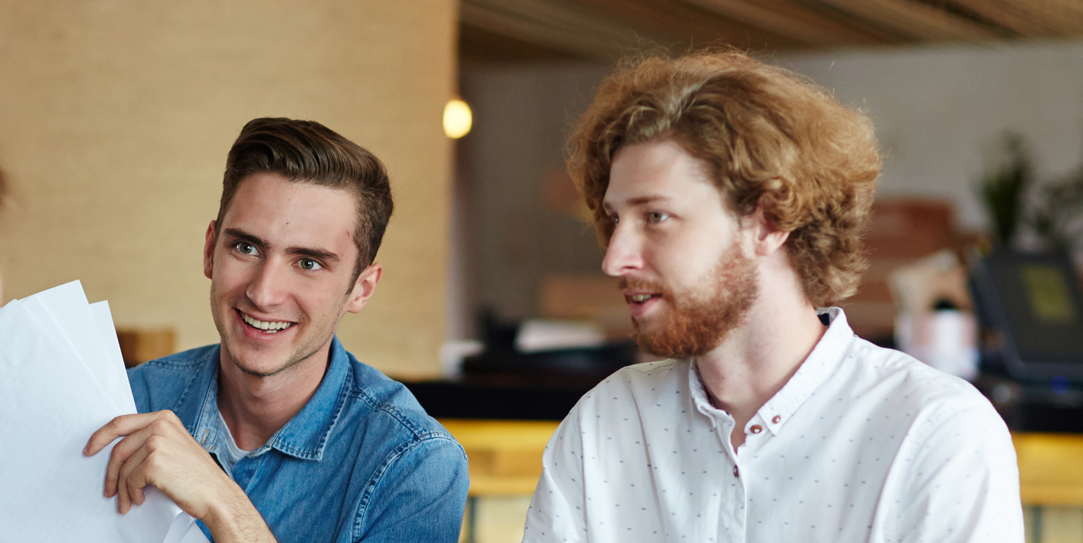 Two men having a discussion, one holding papers and smiling, in a casual indoor setting.