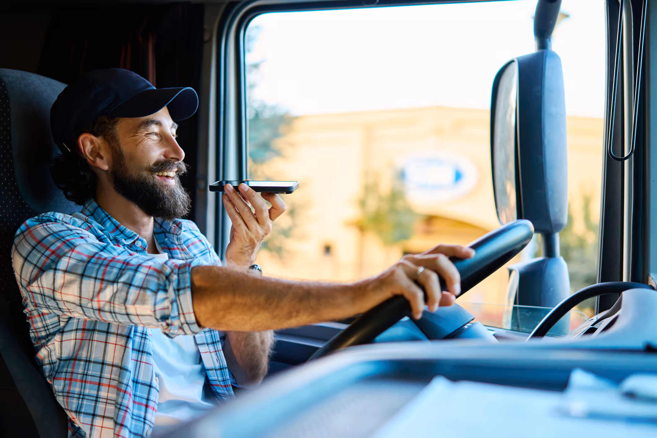 Smiling man driving a truck while speaking into a smartphone held near his mouth.