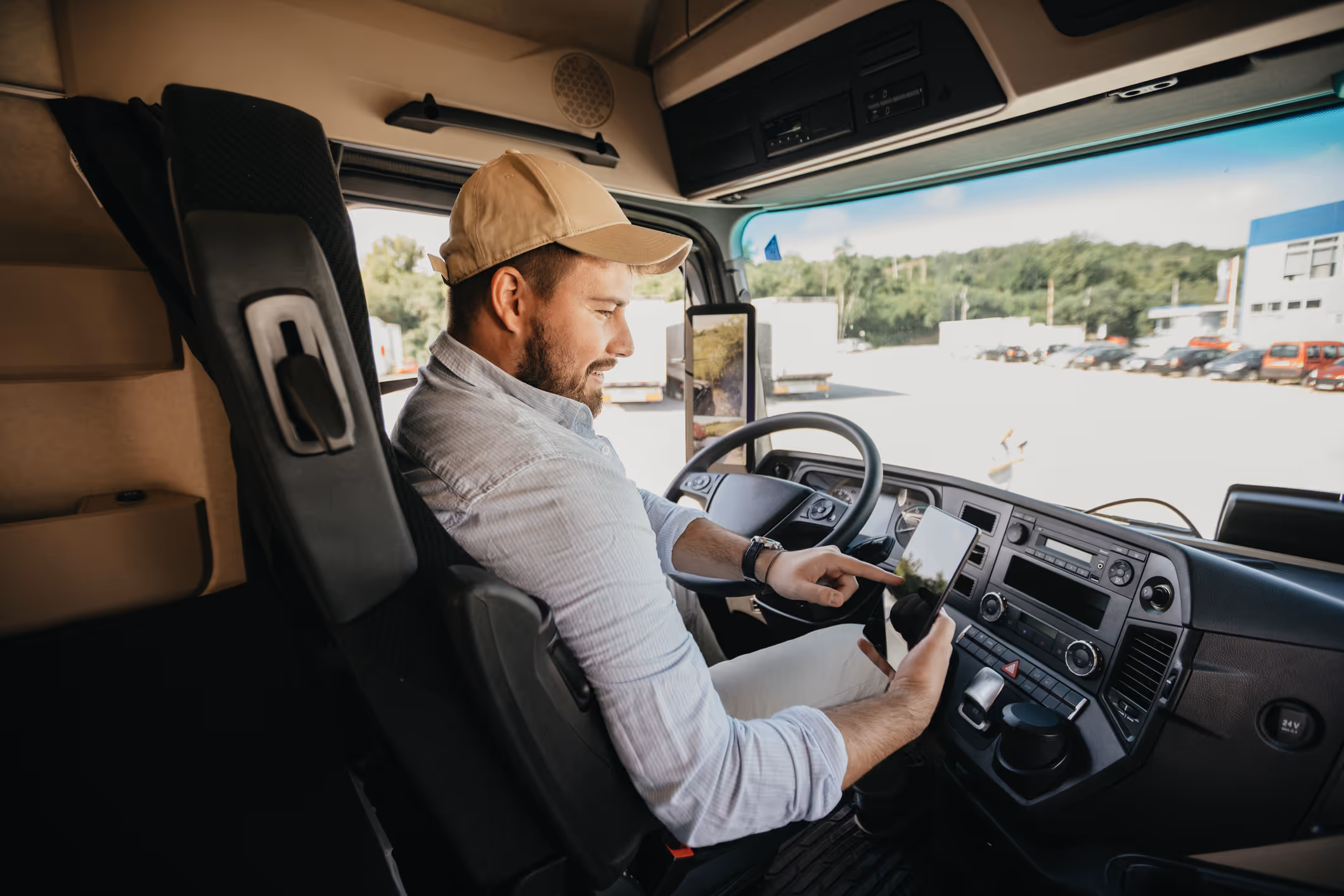 Truck driver sitting in cab using a tablet with a parking lot seen through the windshield.