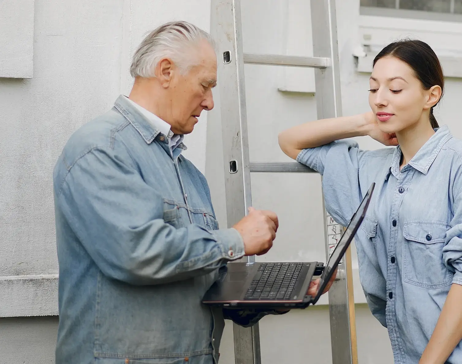 Older man and young woman in denim shirts working with a laptop near a ladder outside a building.