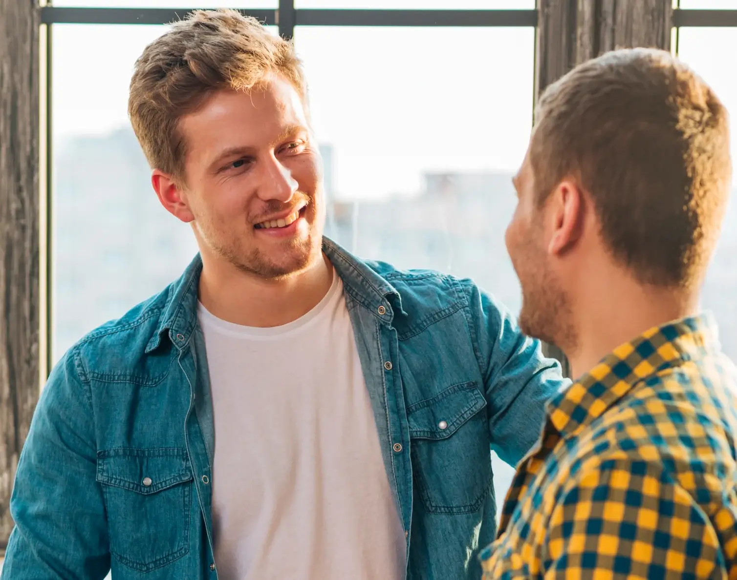 Two men smiling and talking near a window with sunlight shining through.
