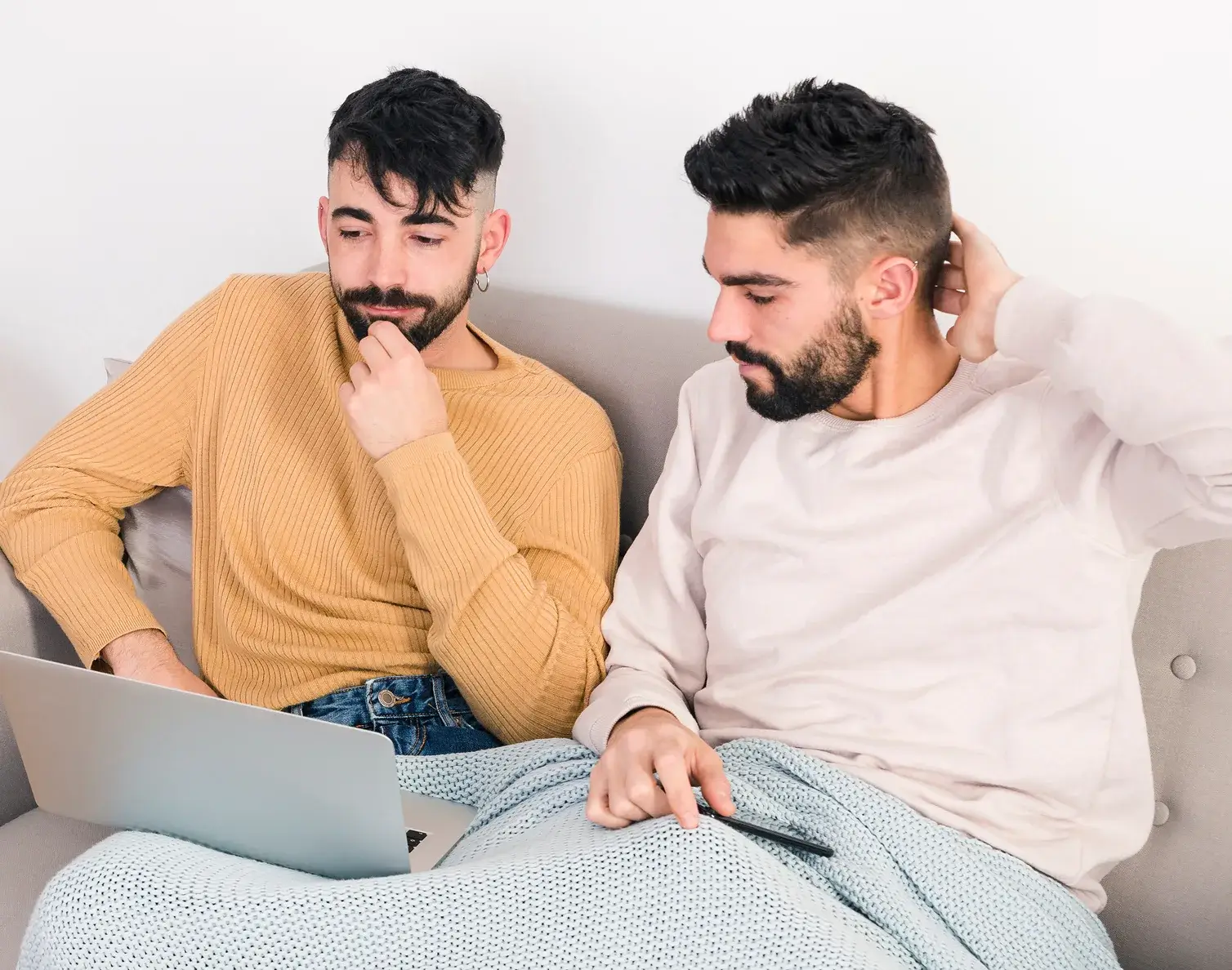 Two men sitting on a couch looking at a laptop and a smartphone with a blanket on their laps.