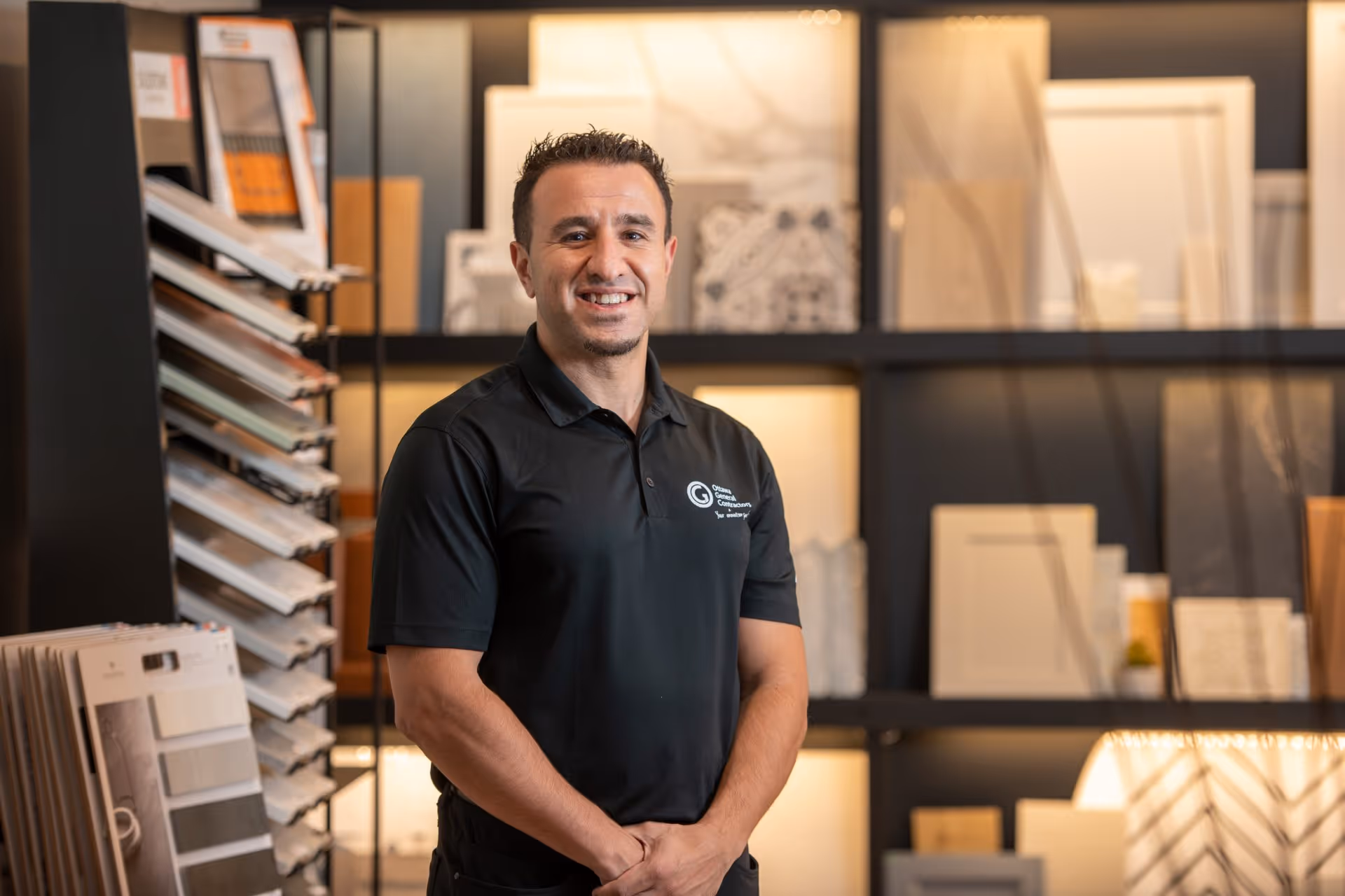 Smiling man in black polo shirt standing in front of tile and countertop samples on display shelves.