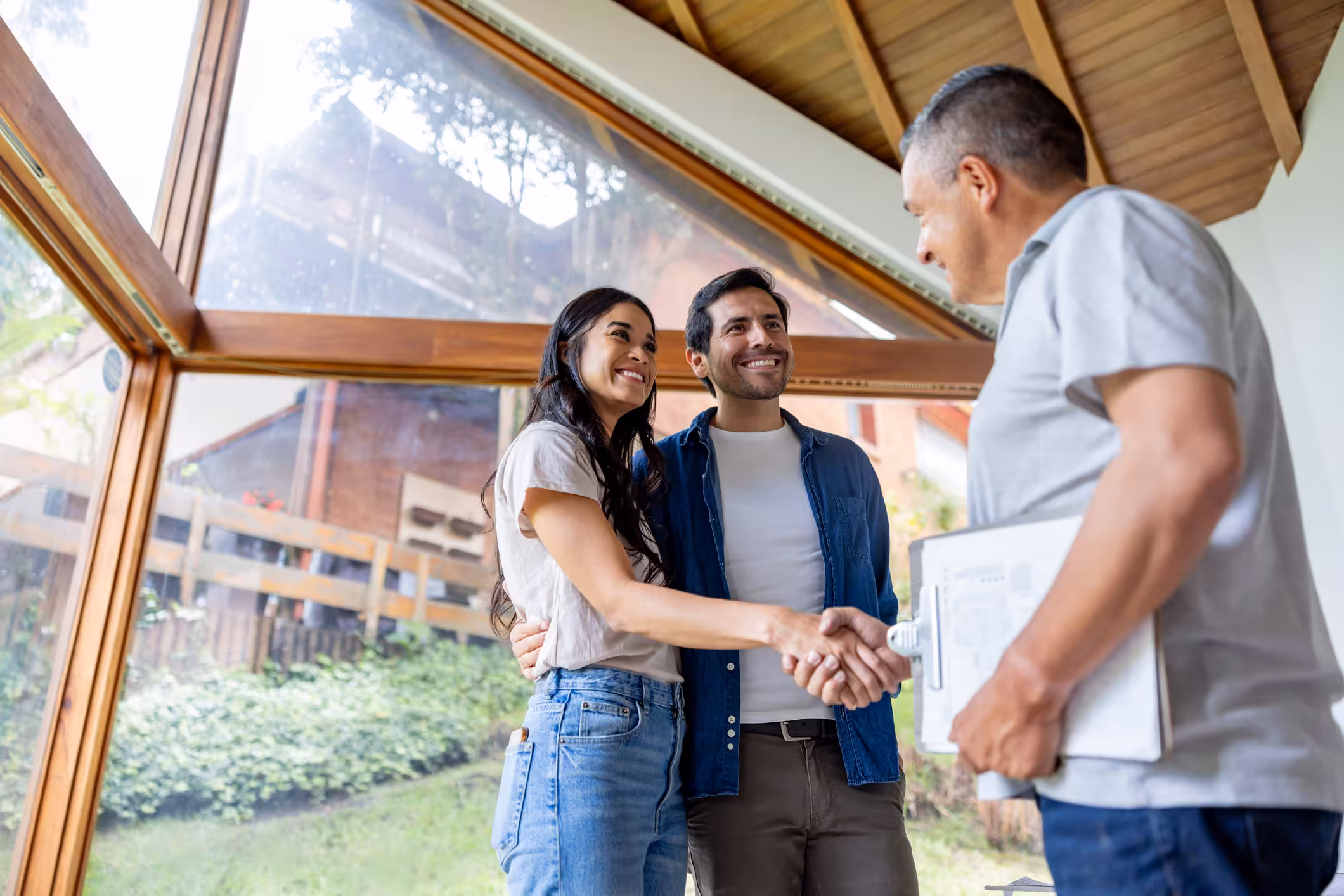 Smiling couple shaking hands with a man holding a clipboard inside a room with large windows.