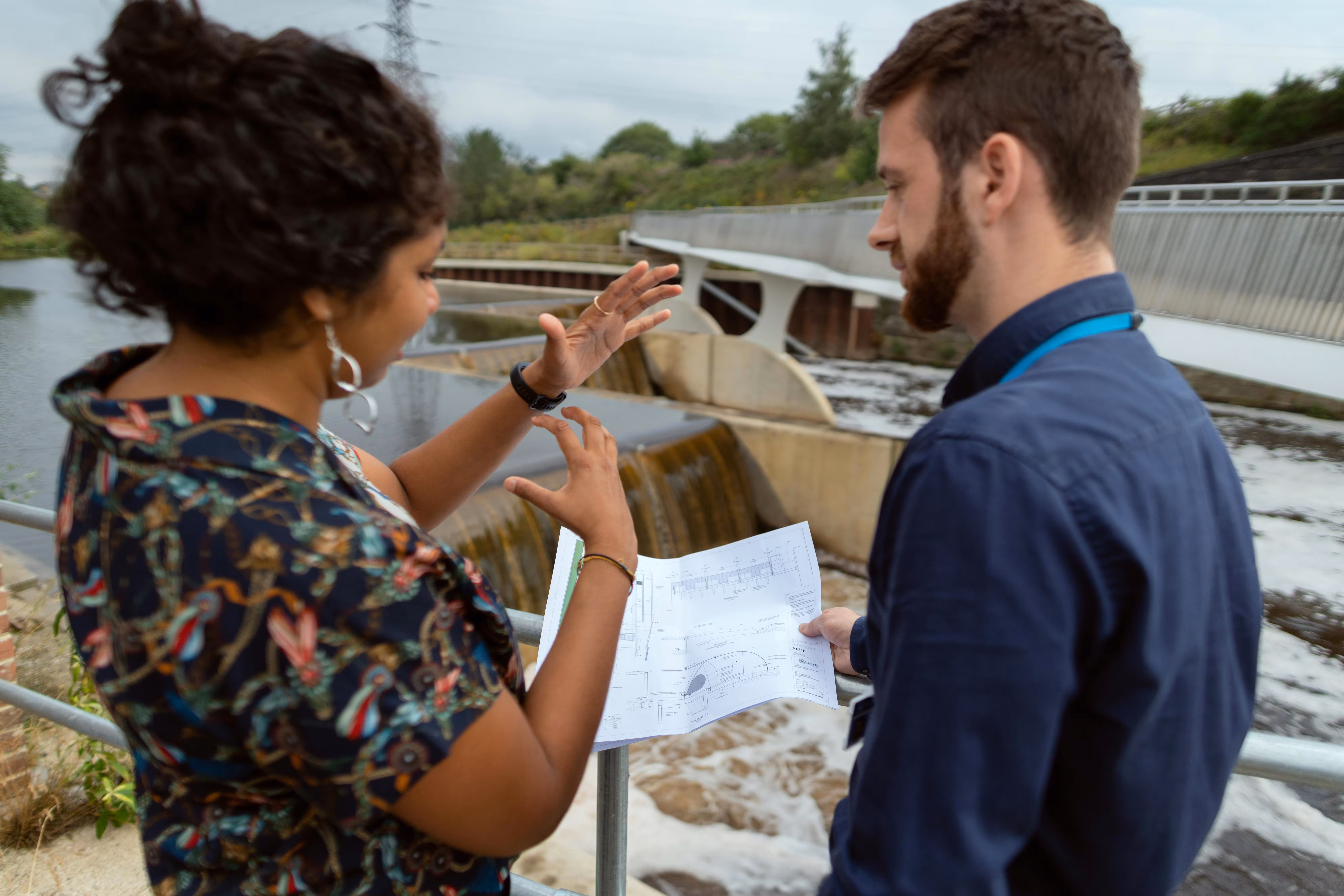 Two engineers discussing plans near a water dam with water flowing below and a bridge in the background.
