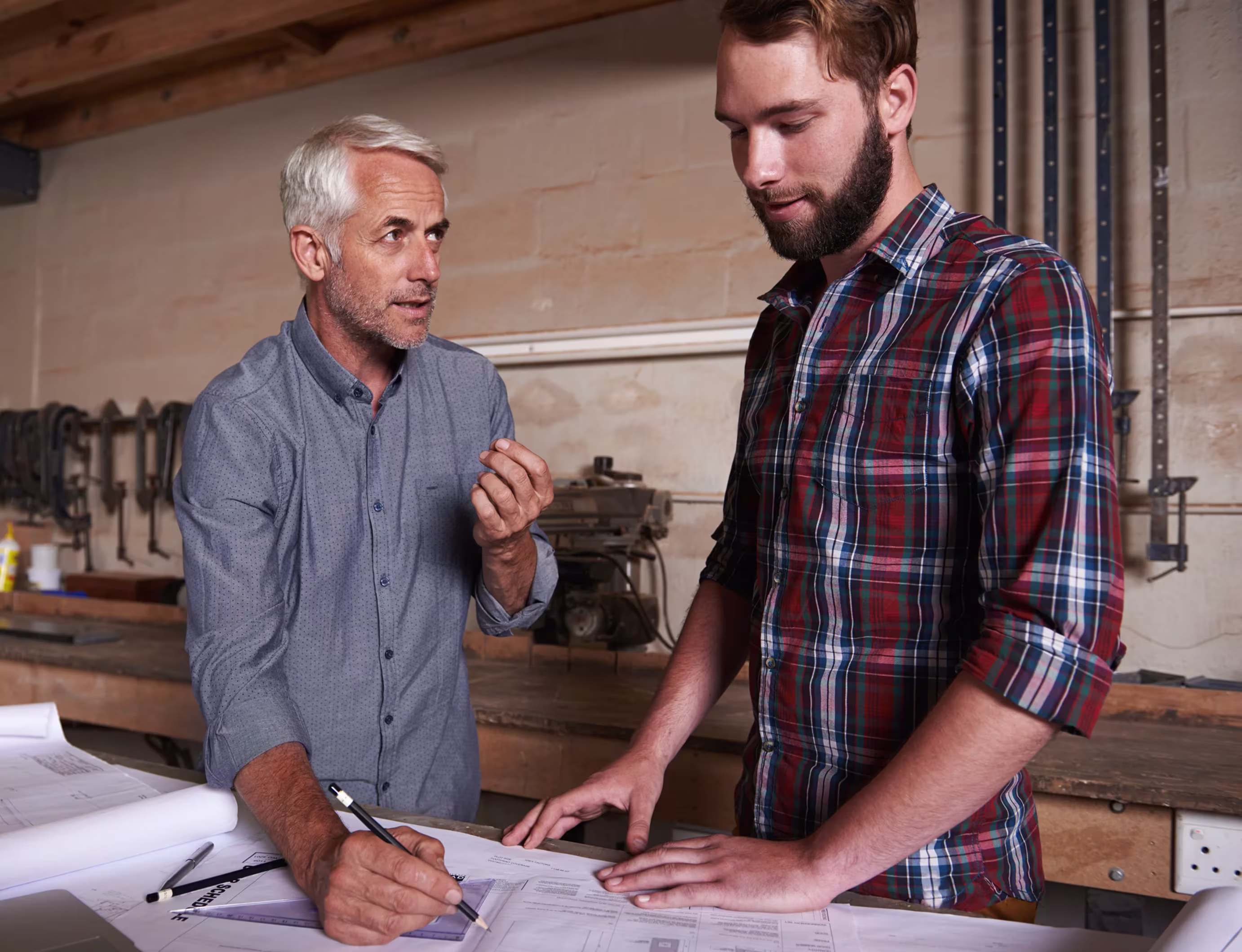 Two men discussing architectural plans on a table in a workshop with tools in the background.