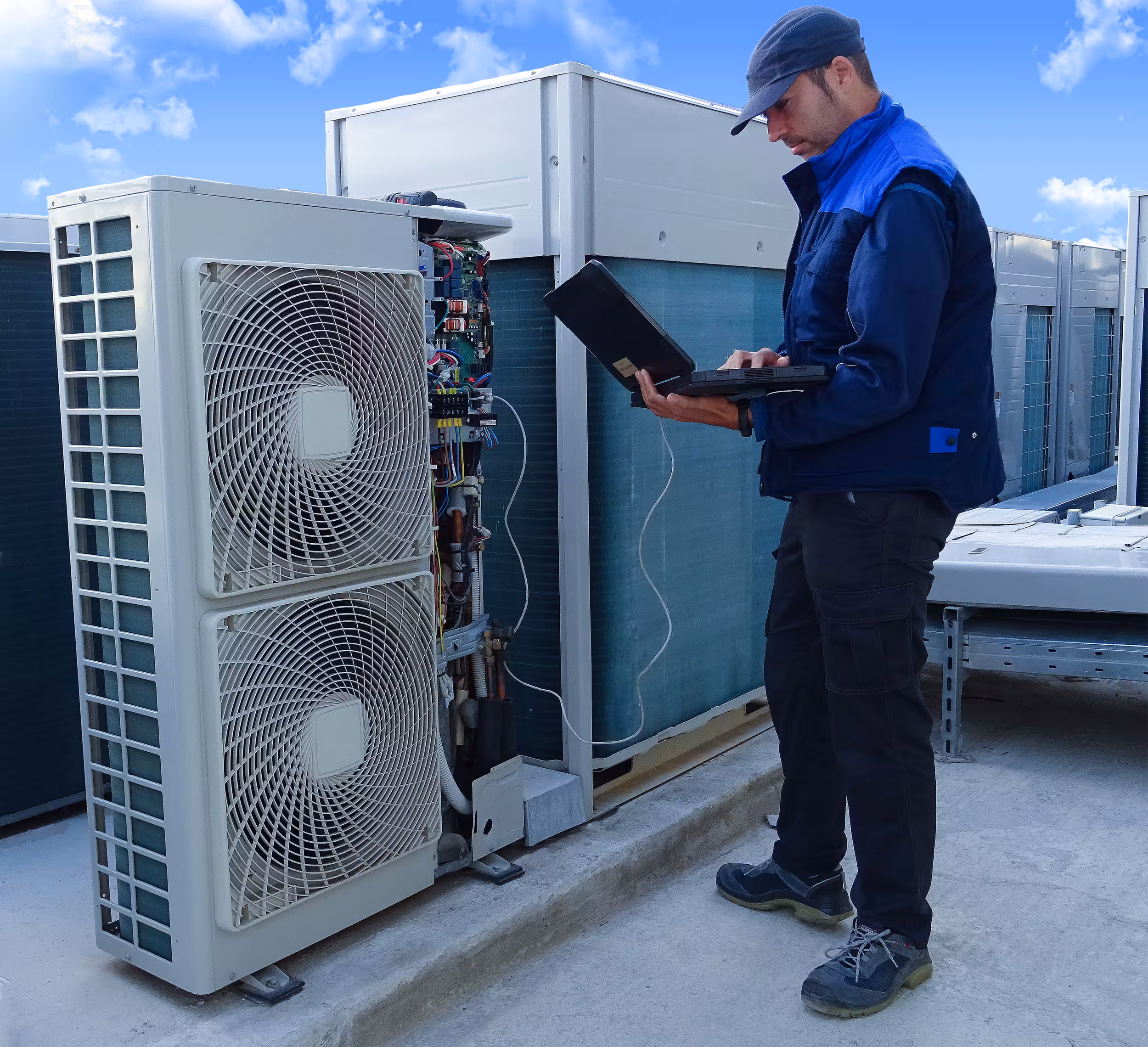 Technician in blue jacket uses a laptop to service an outdoor HVAC unit on a rooftop under blue sky.
